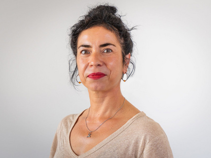 Portraitaufnahme von Severine Klie, sie steht vor weiß/grauem Hintergrund und hat dunkle, nach oben gebundene Haare. Sie trägt roten Lippenstift und eine goldene Kette und hat ein beiges Oberteil an.