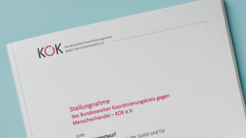 Eine Hand hält ein gedrucktes Dokument des KOK e. V. mit dem Titel Stellungnahme zum Referentenentwurf des Bundesministeriums der Justiz und für Verbraucherschutz zum Gesetz zur Stärkung der strafrechtlichen Verfolgung des Menschenhandels und der sexuellen Ausbeutung. Das Deckblatt zeigt das KOK Logo und formale Angaben, Hintergrund in hellem Blau.