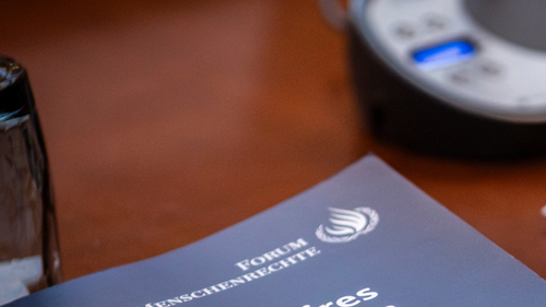 Ein gedrucktes Heft mit dem Titel Aide M&eacute;moires des Forum Menschenrechte 2025 liegt auf einem Tisch. Daneben stehen ein Glas und ein technisches Ger&auml;t.