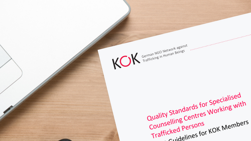 Schr&auml;g liegendes Deckblatt einer englischsprachigen KOK-Publikation auf einem Holztisch. Titel &bdquo;Quality Standards for Specialised Counselling Centres Working with Trafficked Persons&ldquo;. Daneben liegen ein Smartphone und ein Laptop.