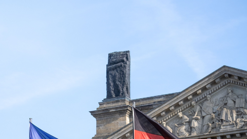 Vor dem Reichstagsgebäude in Berlin wehen die Flaggen der Europäischen Union und Deutschlands auf zwei Fahnenmasten. Im Hintergrund sind die Säulen und der Giebel des Gebäudes zu sehen. Das Bild steht sinnbildlich für die europäische und nationale Gesetzgebung sowie die Zusammenarbeit zwischen Deutschland und der Europäischen Union.