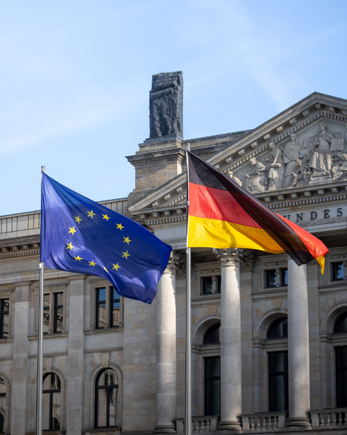 Vor dem Reichstagsgebäude in Berlin wehen die Flaggen der Europäischen Union und Deutschlands auf zwei Fahnenmasten. Im Hintergrund sind die Säulen und der Giebel des Gebäudes zu sehen. Das Bild steht sinnbildlich für die europäische und nationale Gesetzgebung sowie die Zusammenarbeit zwischen Deutschland und der Europäischen Union.