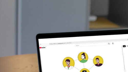 Ein aufgeklappter Laptop steht auf einem Holztisch neben einer Tasse. Auf dem Bildschirm ist ein YouTube-Video zu sehen, das eine Grafik mit vier verbundenen Personen-Illustrationen zeigt, die ein Netzwerk darstellen.