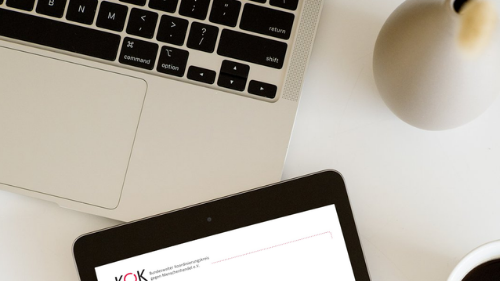 Draufsicht auf einen Schreibtisch mit Laptop, Kaffeetasse und Tablet. Auf dem Tablet ist ein Dokument mit der Überschrift KOK-Rechtsprechungsdatenbank zu sehen.