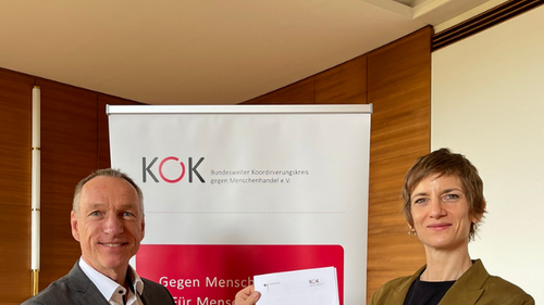 Sophia Wirsching, Geschäftsführerin des KOK, und Markus Koths, Leiter der Abteilung Schwere und Organisierte Kriminalität beim Bundeskriminalamt (BKA), stehen vor einem Roll-up des KOK. Sie schütteln sich die Hand und halten gemeinsam die Kooperatiosvereinbarung in die Kamera.
