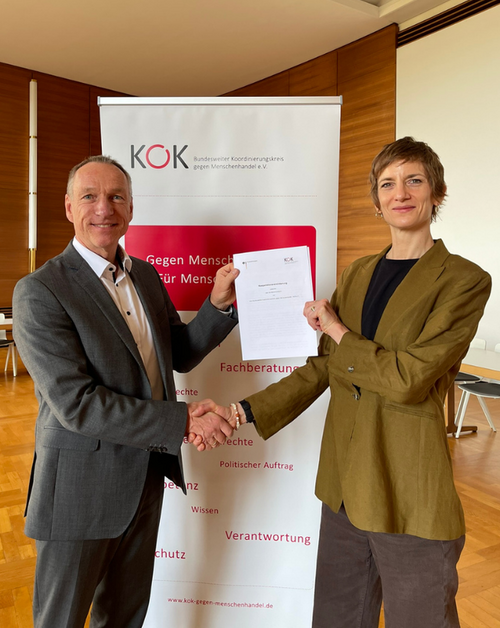 Sophia Wirsching, Gesch&auml;ftsf&uuml;hrerin des KOK, und Markus Koths, Leiter der Abteilung Schwere und Organisierte Kriminalit&auml;t beim Bundeskriminalamt (BKA), stehen vor einem Roll-up des KOK. Sie sch&uuml;tteln sich die Hand und halten gemeinsam die Kooperatiosvereinbarung in die Kamera.