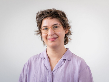 Portraitaufnahme von Charlotte Kunath. Sie steht vor einer grau-weißen Wand und hat eine lilane Bluse an. Sie hat braune, leicht gewellte kurze Haare und trägt eine Brille.