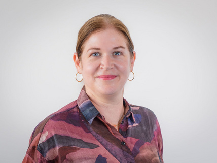 Portraitaufnahme von Sarah Schwarze: Sie steht vor einer weiß-grauen Wand, hat hellbraune Haare und trägt einen Pferdeschwanz. Sie hat eine pink, lila gemusterte Bluse an, trägt Ohrringe und rosafarbenen Lippenstift