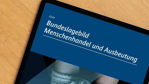 Auf einem Schreibtisch liegt ein Tablet, auf dem das Bundeslagebild Menschenhandel und Ausbeutung geöffnet ist. Man sieht das Cover: Titel "Bundeslagebild Menschenhandel und Ausbeutung 2024) steht im oberen Bereich auf einem blauen Feld. Darunter sind zwei gefesselte Hände zu sehen. Die Nägel der Hände sind rot lackiert. Neben dem Tablet sind Zettel und Stift zu sehen.