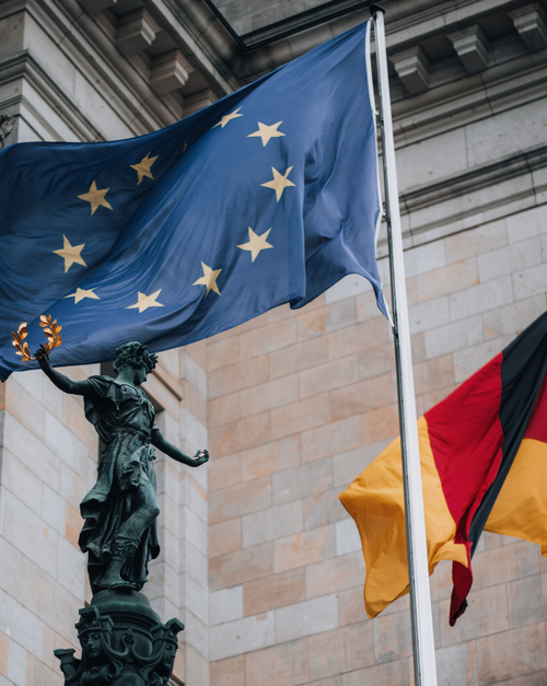 Nahaufnahme einer wehenden Flagge der Europ&auml;ischen Union vor der Sandsteinfassade des Reichstagsgeb&auml;udes in Berlin. Im Vordergrund steht eine Bronzestatue der G&ouml;ttin Viktoria mit einem Lorbeerzweig in der erhobenen Hand. Rechts ist teilweise die deutsche Flagge zu sehen. 