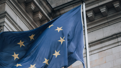 Nahaufnahme einer wehenden Flagge der Europäischen Union vor der Sandsteinfassade des Reichstagsgebäudes in Berlin. Im Vordergrund steht eine Bronzestatue der Göttin Viktoria mit einem Lorbeerzweig in der erhobenen Hand. Rechts ist teilweise die deutsche Flagge zu sehen.