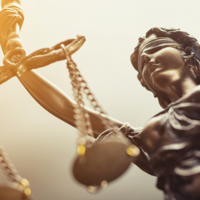 Nahaufnahme einer Statue der Justitia. Die Figur hält eine Waage in der Hand, trägt eine Augenbinde und ist in warmes Licht getaucht. Die Aufnahme ist leicht von unten fotografiert, sodass die Skulptur imposant wirkt.