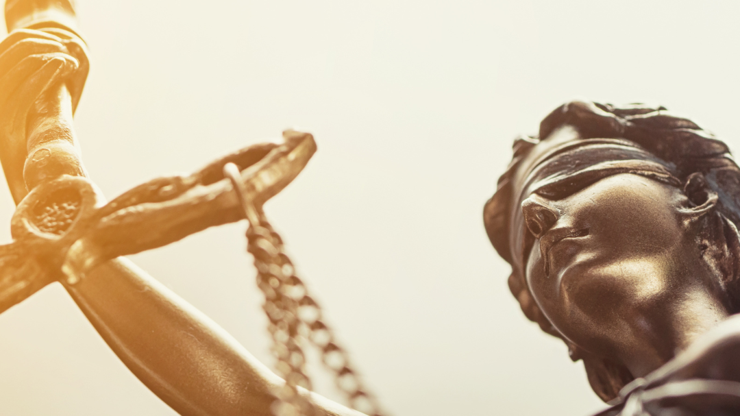 Nahaufnahme einer Statue der Justitia. Die Figur hält eine Waage in der Hand, trägt eine Augenbinde und ist in warmes Licht getaucht. Die Aufnahme ist leicht von unten fotografiert, sodass die Skulptur imposant wirkt.