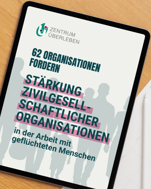 Ein Tablet auf einem Holzschreibtisch zeigt das Titelblatt einer Publikation des Zentrum &Uuml;berleben mit der &Uuml;berschrift &bdquo;62 Organisationen fordern St&auml;rkung zivilgesellschaftlicher Organisationen in der Arbeit mit gefl&uuml;chteten Menschen&ldquo;. Im Hintergrund des Titelblatts sind stilisierte Silhouetten von mehreren Menschen abgebildet, die Hand in Hand nebeneinander stehen.