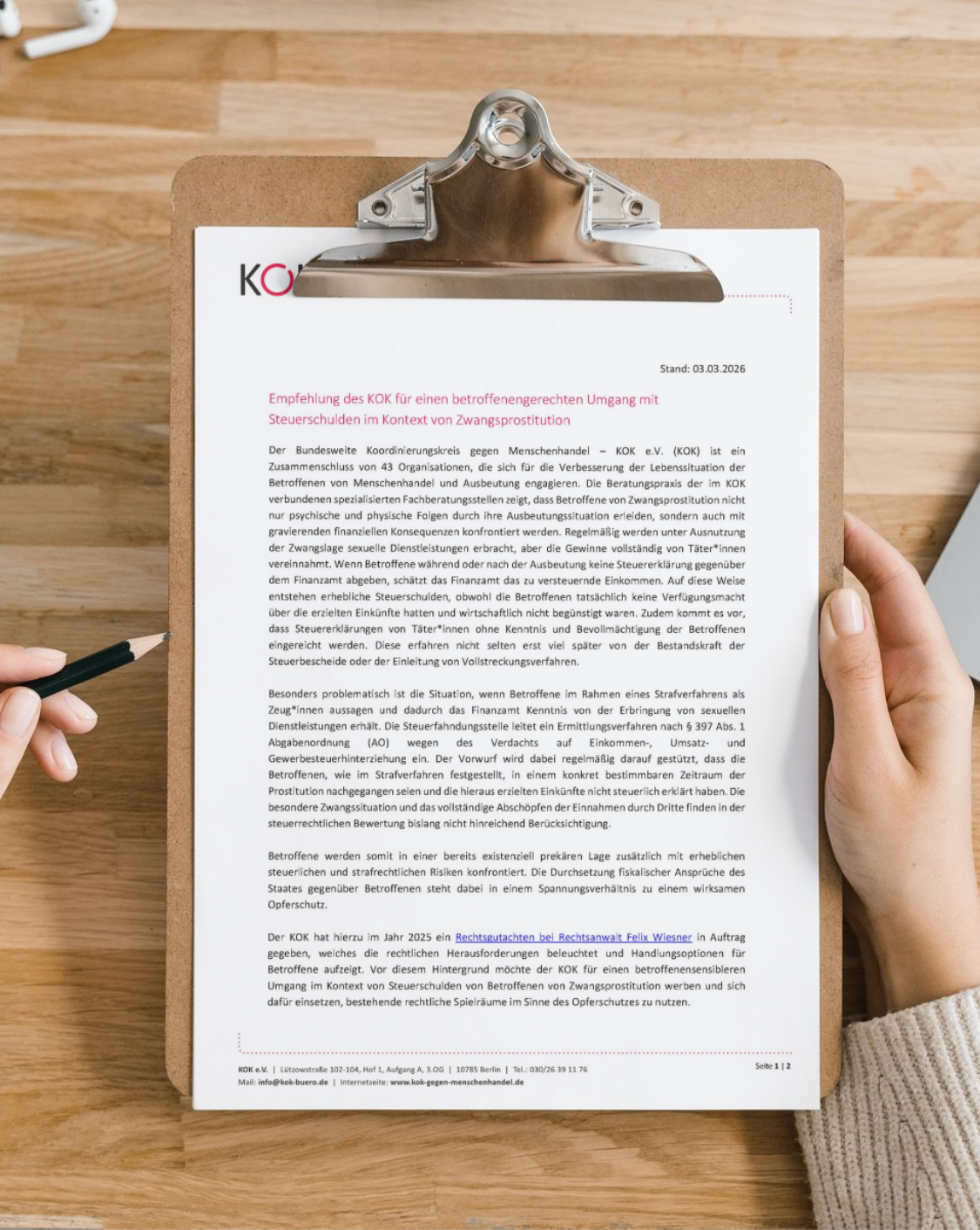 Person h&auml;lt ein Klemmbrett mit einem deutschsprachigen Informationspapier des Bundesweiten Koordinierungskreises gegen Menschenhandel (KOK e. V.) auf einem Holztisch, Titel: Empfehlung des KOK e.V. f&uuml;r einen betroffenengerechten Umgang mit Steuerschulden im Kontext von Zwangsprostitution