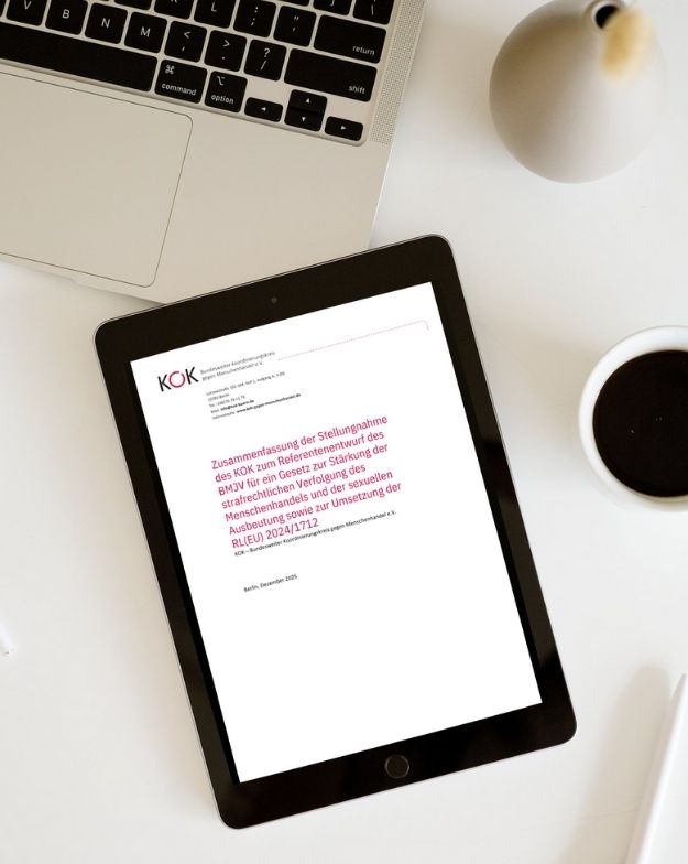 Tablet auf einem Schreibtisch mit ge&ouml;ffneter KOK-Publikation. Zu sehen ist das Deckblatt einer Stellungnahme zur Reform der strafrechtlichen Verfolgung von Menschenhandel und sexueller Ausbeutung. Daneben liegen ein Laptop und eine Tasse Kaffee.