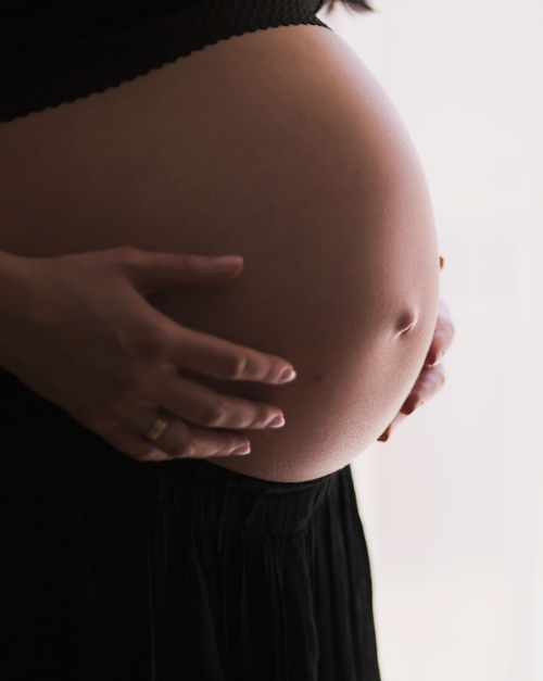 Eine schwangere Person steht vor einem hellen Hintergrund und h&auml;lt sich den Bauch. Die Person tr&auml;gt dunkle Kleidung.