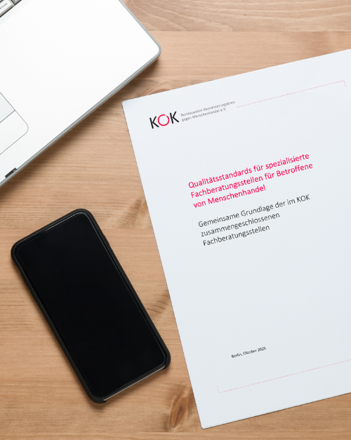 Ein Smartphone und ein Laptop liegen auf einem Holztisch. Daneben liegt ein Dokument mit dem Titel „Qualitätsstandards für spezialisierte Fachberatungsstellen für Betroffene von Menschenhandel“ vom KOK – Bundesweiter Koordinierungskreis gegen Menschenhandel e.V.
