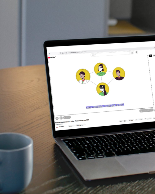 Ein aufgeklappter Laptop steht auf einem Holztisch neben einer Tasse. Auf dem Bildschirm ist ein YouTube-Video zu sehen, das eine Grafik mit vier verbundenen Personen-Illustrationen zeigt, die ein Netzwerk darstellen.