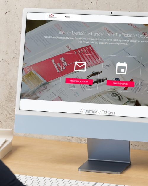 Alt-Text: Desktop-Monitor auf einem Schreibtisch. Auf dem Bildschirm die Startseite der Online Anlaufstelle Menschenhandel des KOK. Überschrift Hilfe bei Menschenhandel. Zwei große Schaltflächen mit Symbolen für E Mail Anfrage stellen und Termin buchen. Im Hintergrund liegen Informationsmaterialien und Flyer.