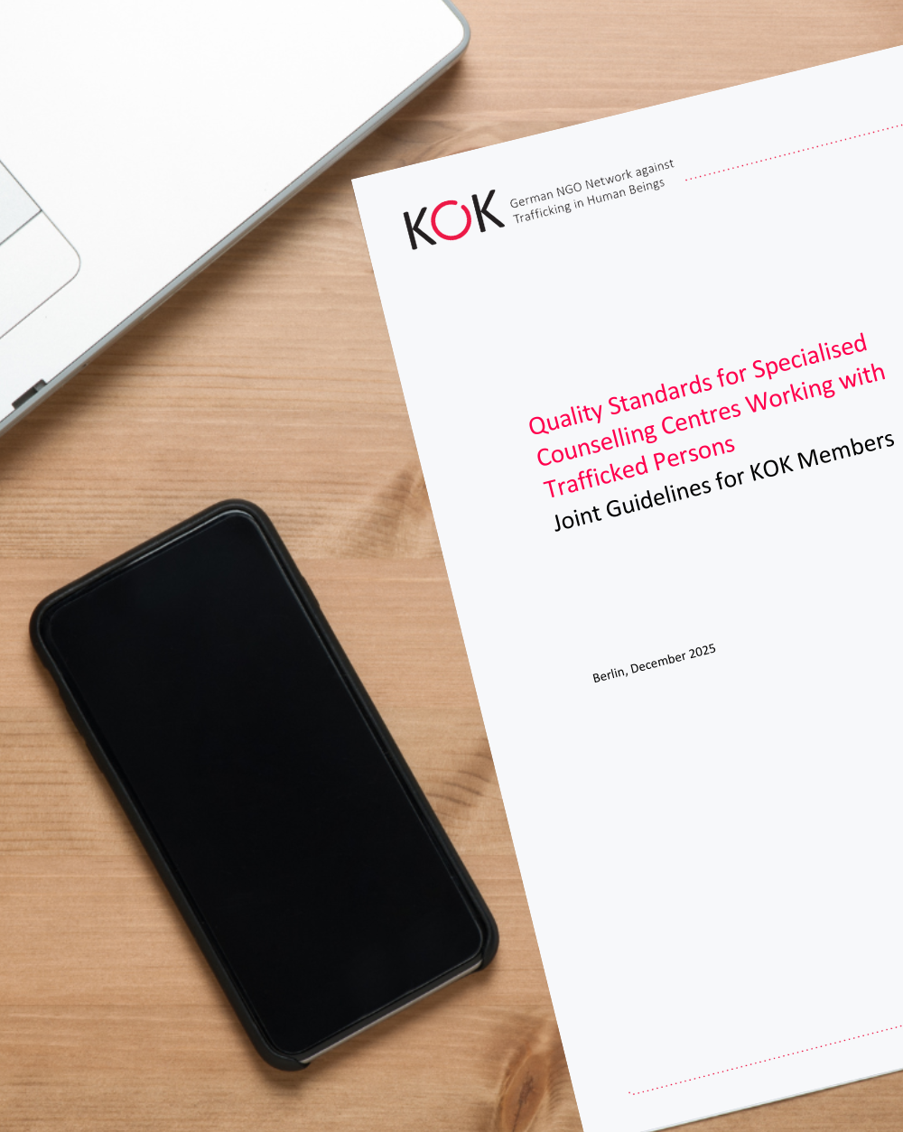 Schr&auml;g liegendes Deckblatt einer englischsprachigen KOK-Publikation auf einem Holztisch. Titel &bdquo;Quality Standards for Specialised Counselling Centres Working with Trafficked Persons&ldquo;. Daneben liegen ein Smartphone und ein Laptop.