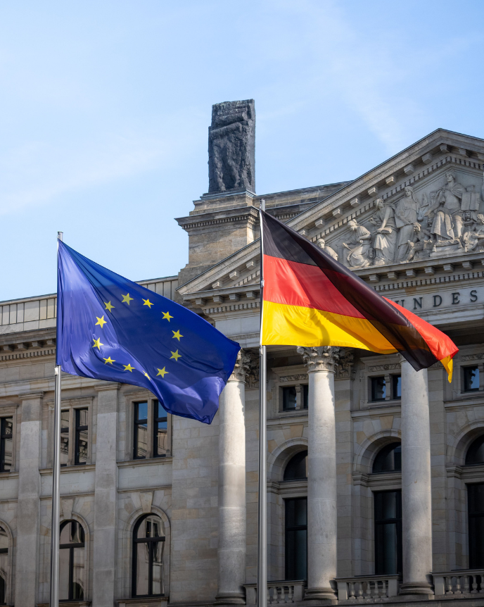 Vor dem Reichstagsgeb&auml;ude in Berlin wehen die Flaggen der Europ&auml;ischen Union und Deutschlands auf zwei Fahnenmasten. Im Hintergrund sind die S&auml;ulen und der Giebel des Geb&auml;udes zu sehen. Das Bild steht sinnbildlich f&uuml;r die europ&auml;ische und nationale Gesetzgebung sowie die Zusammenarbeit zwischen Deutschland und der Europ&auml;ischen Union.